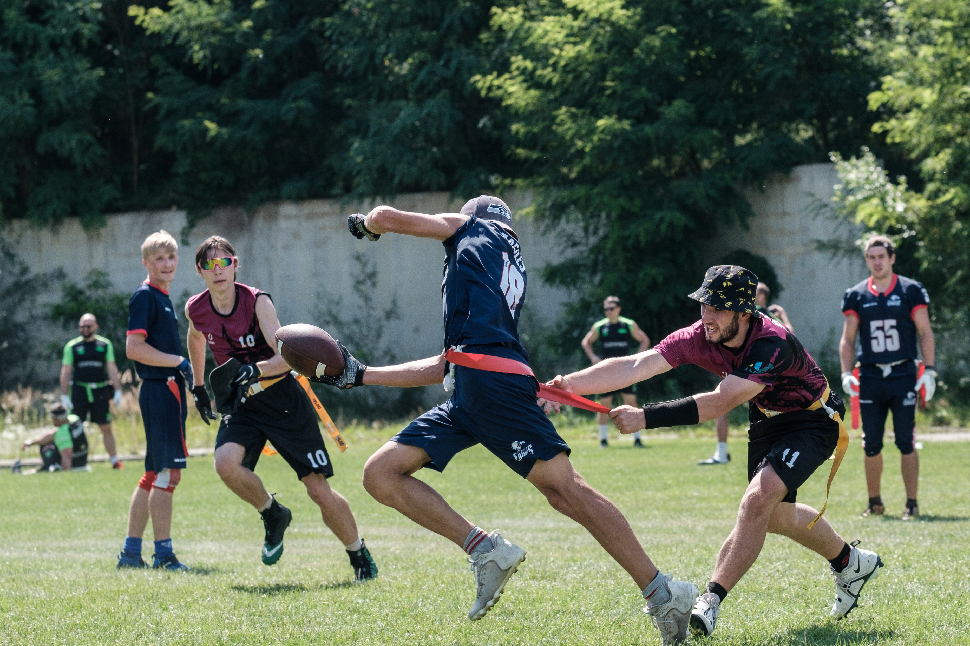 A group of young people playing flag football outdoors, one player in a blue jersey lunges to avoid being flagged by another in a maroon jersey. Forest in the background