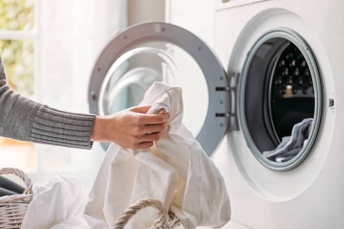 A person loading a white sheet into the washing machine from a basket. Image is likely related to household chores or laundry tips