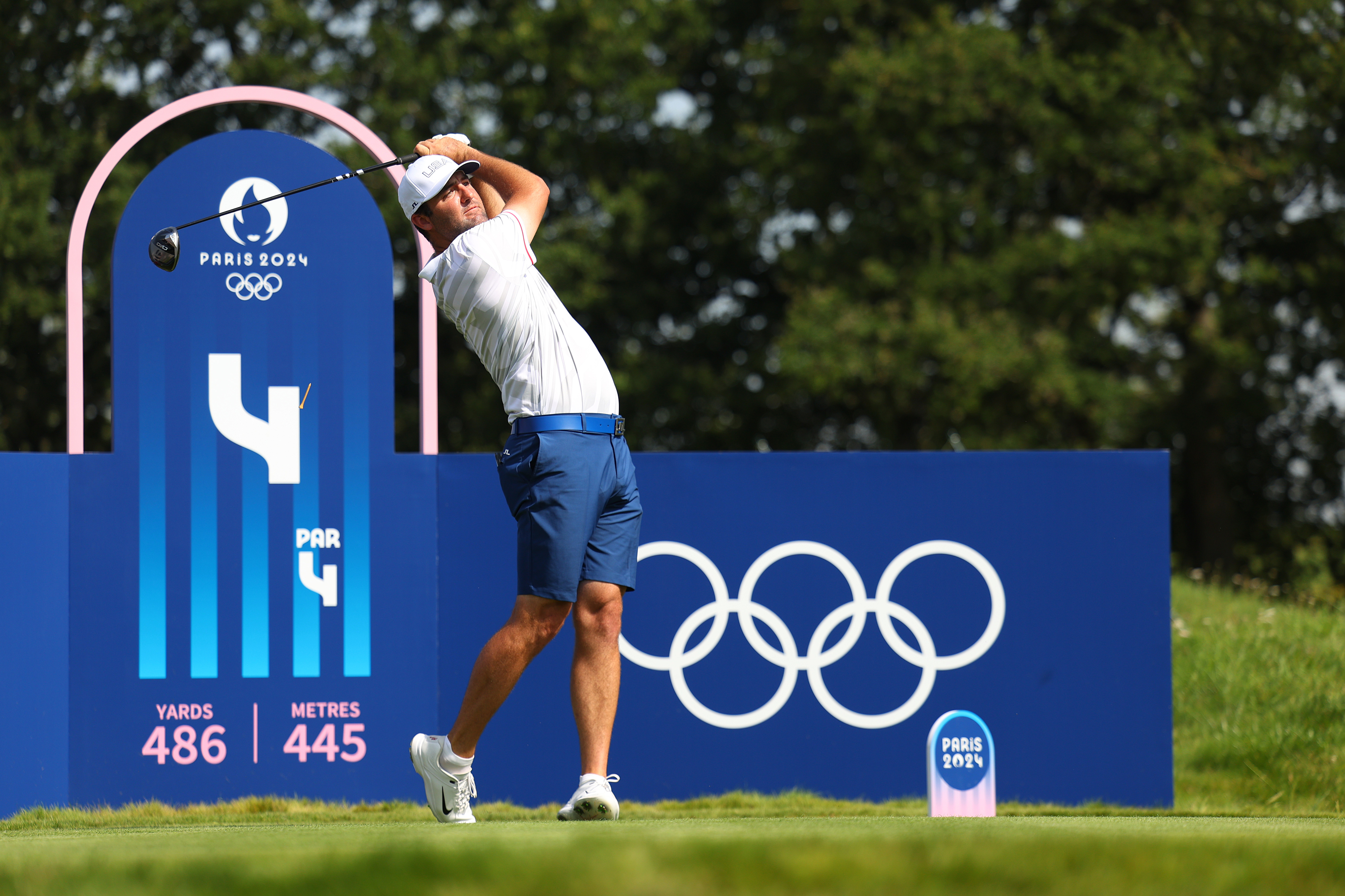 Golfer Alexander Noren swinging a club on a course during the Paris 2024 Olympic Games. A sign shows hole details: 4, 486 yards, 445 meters, par 4