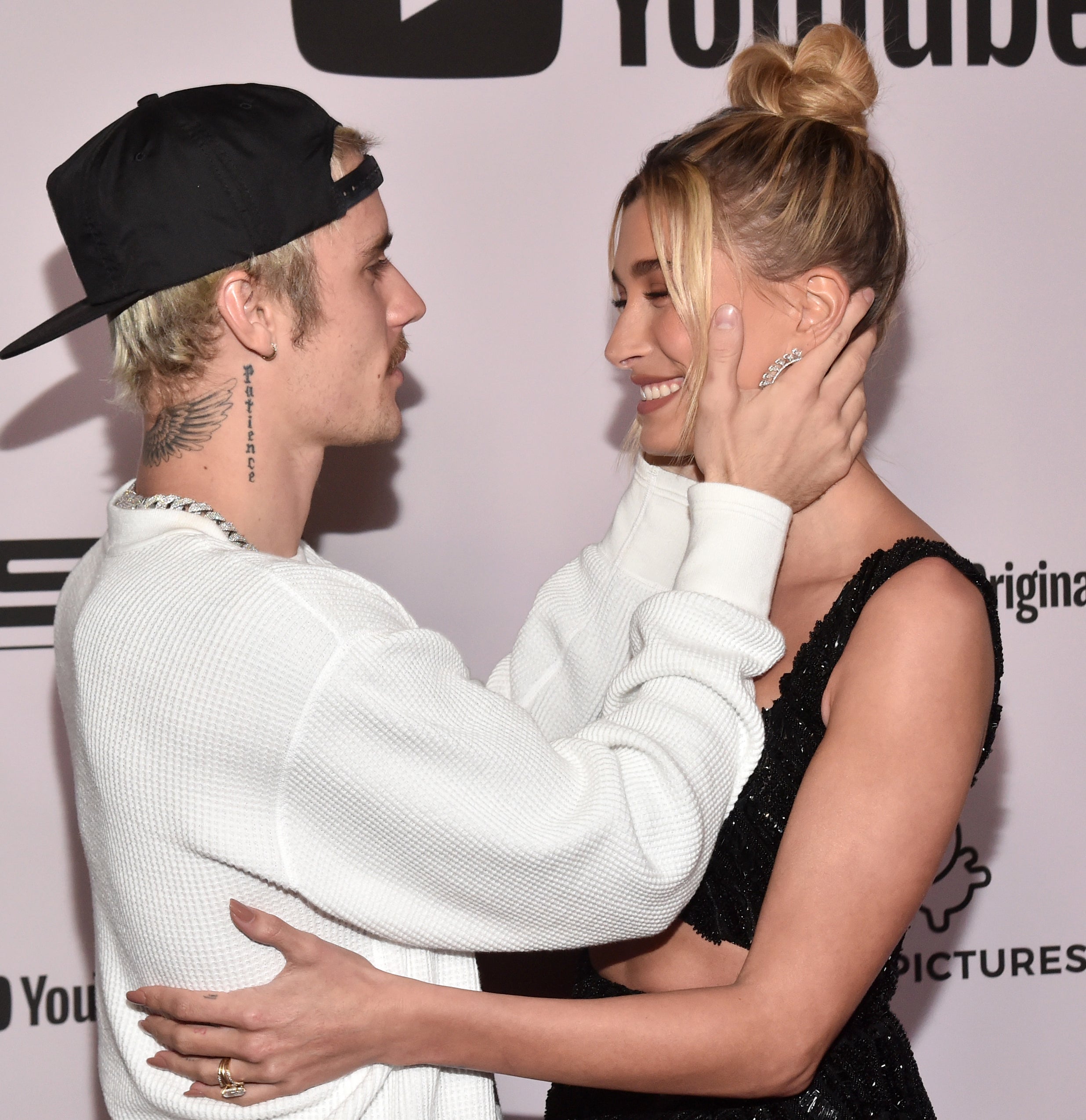 Justin Bieber, wearing a white long-sleeve shirt and a black cap, cups Hailey Bieber&#x27;s face, who is in a black dress, on a YouTube Originals red carpet