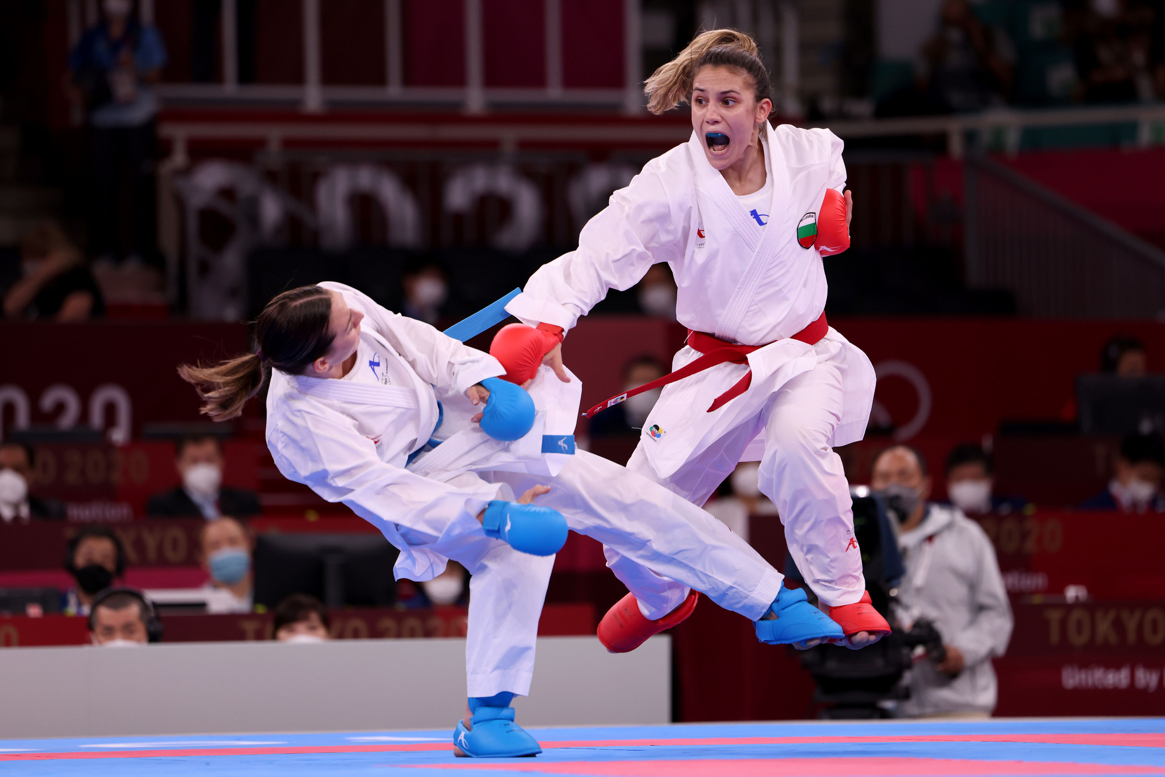 Two martial artists in white uniforms and protective gear spar during an Olympic karate match, one airborne delivering a kick. Audience visible in the background