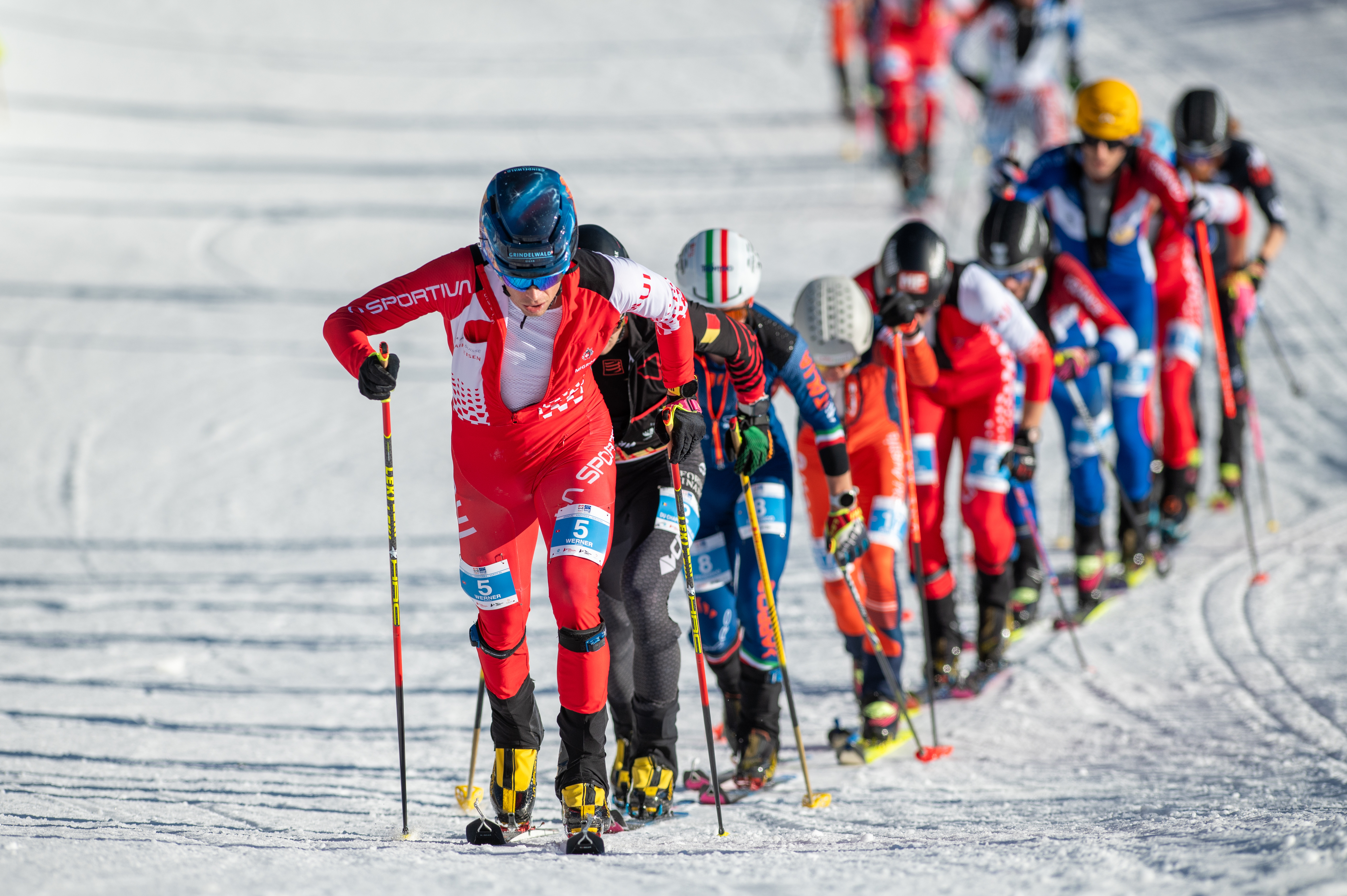 A group of skiers compete in a cross-country skiing race, skiing uphill in a line on a snowy terrain