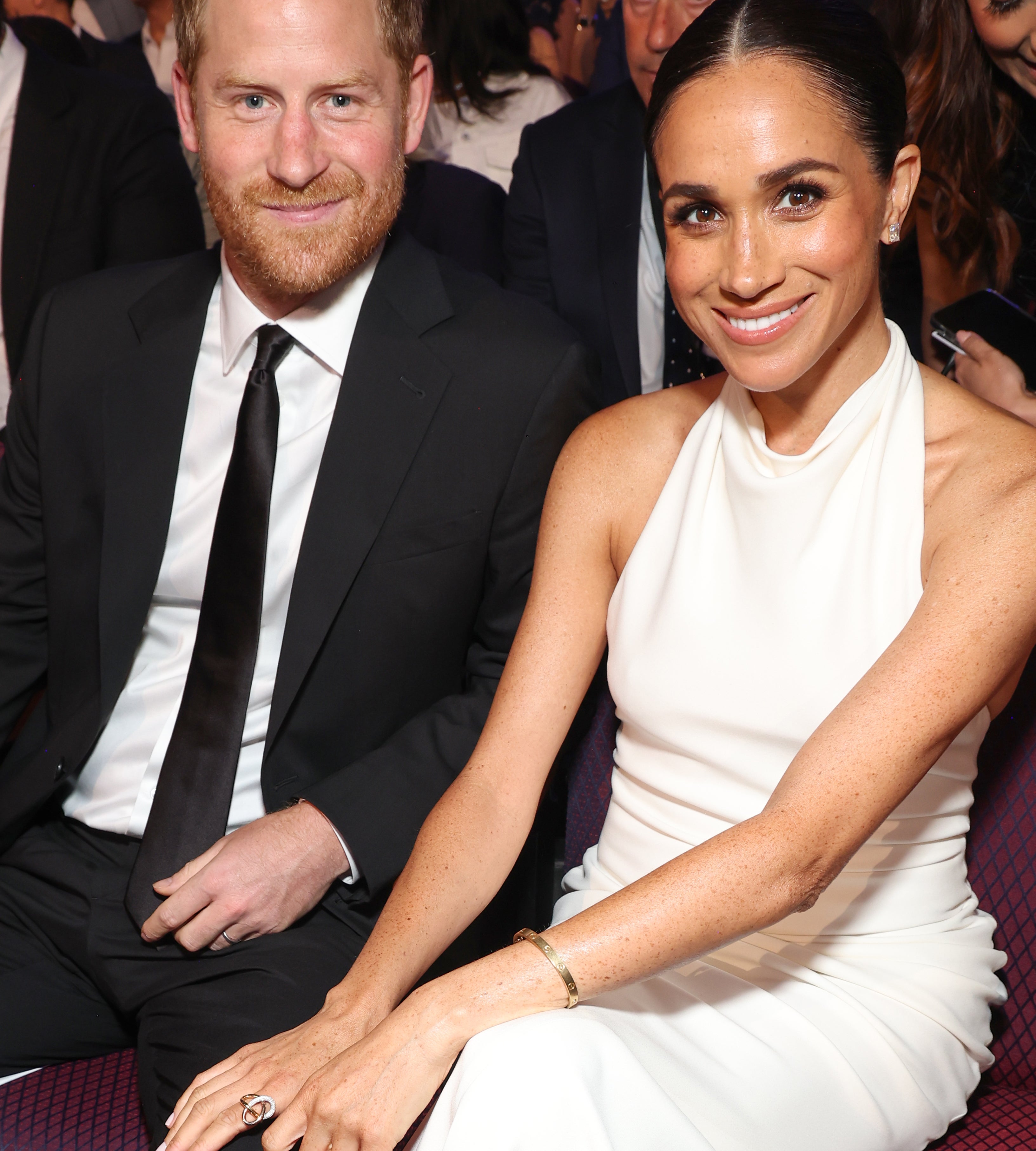 Prince Harry and Meghan Markle sitting together at an event. He is wearing a black suit and tie; she is wearing a white sleeveless dress