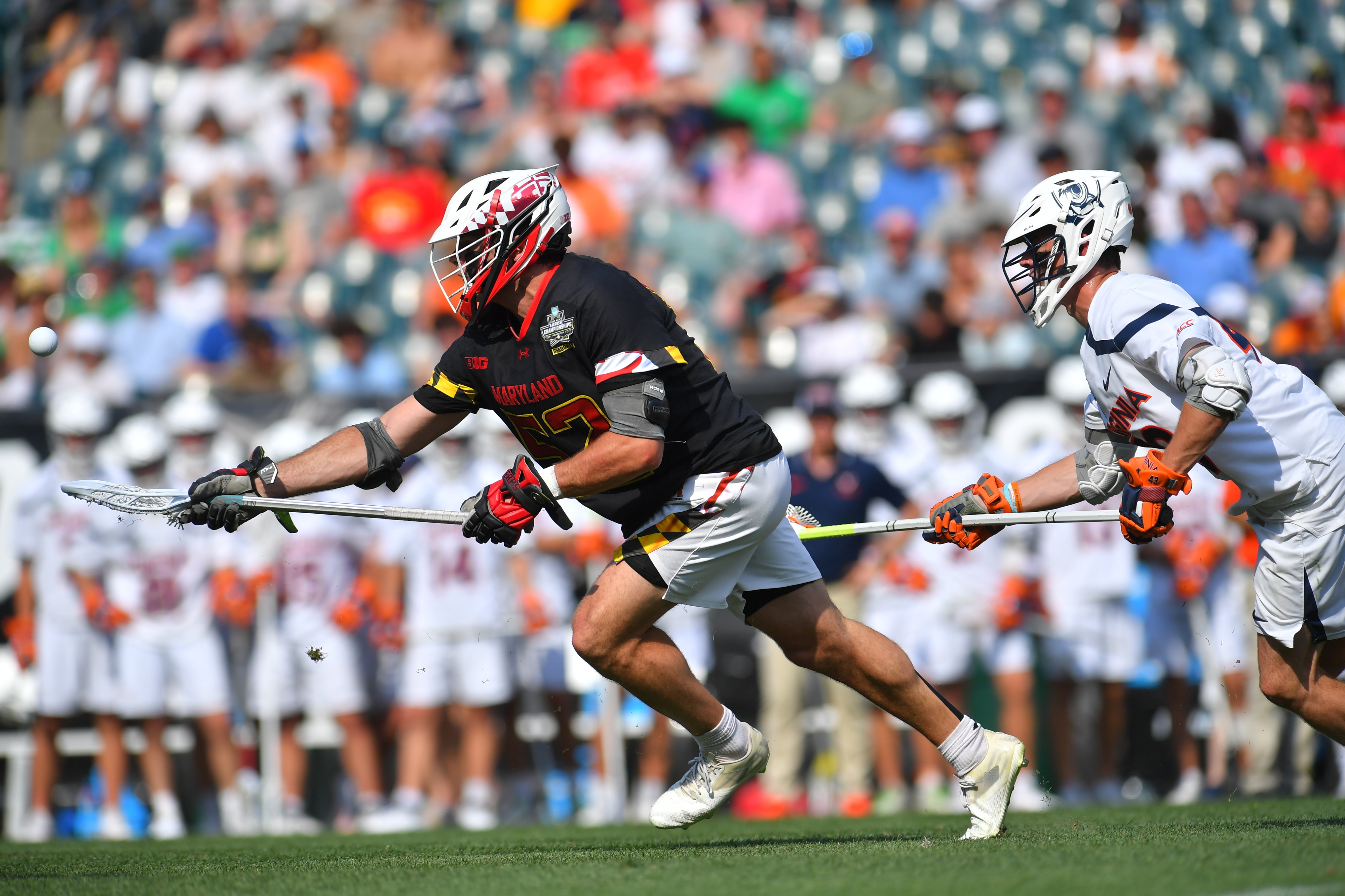 Two lacrosse players, one from the University of Maryland, compete during a game. The Maryland player is running with the ball, while the opponent closely follows