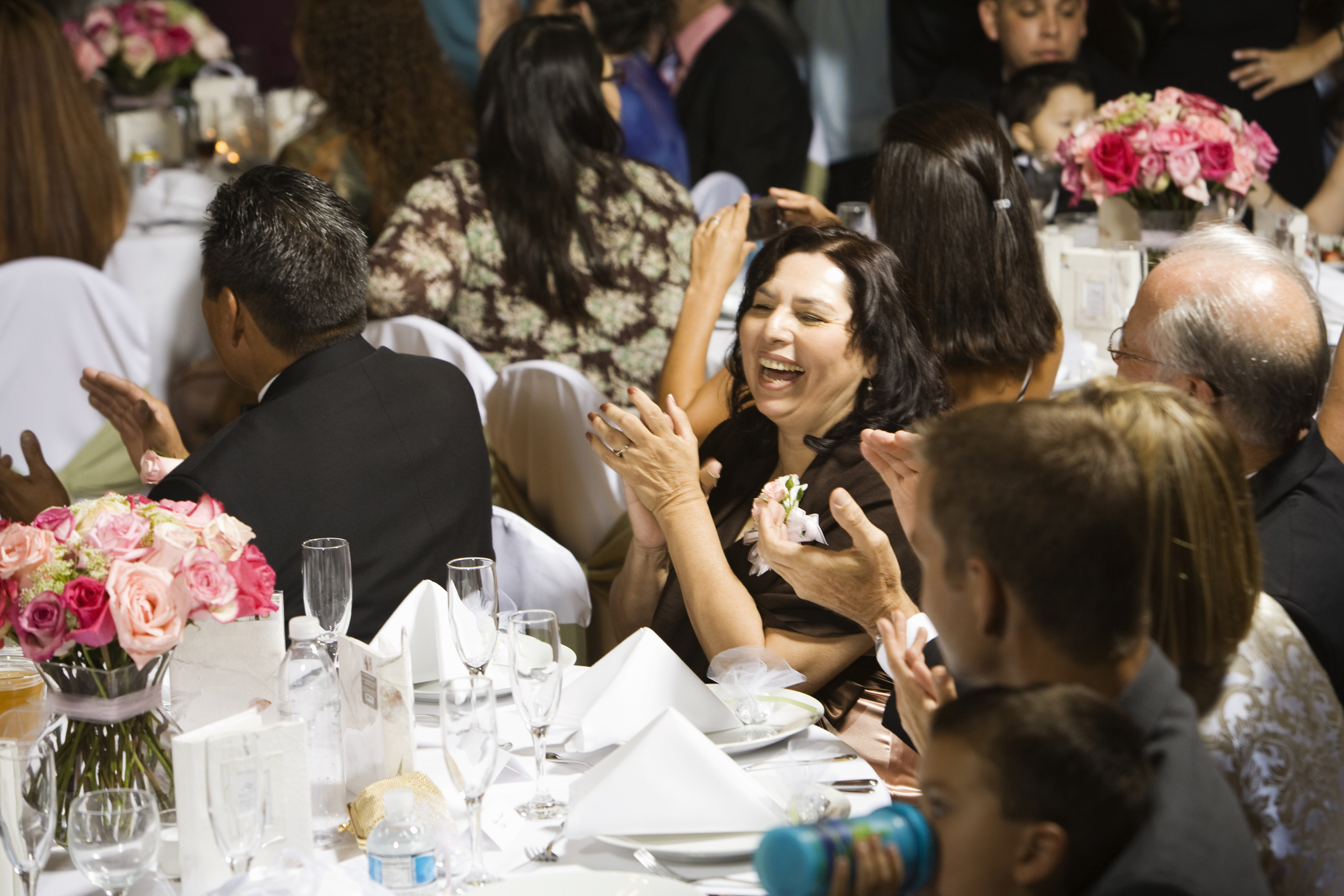 Guests seated at a wedding reception table, smiling, talking, and clapping