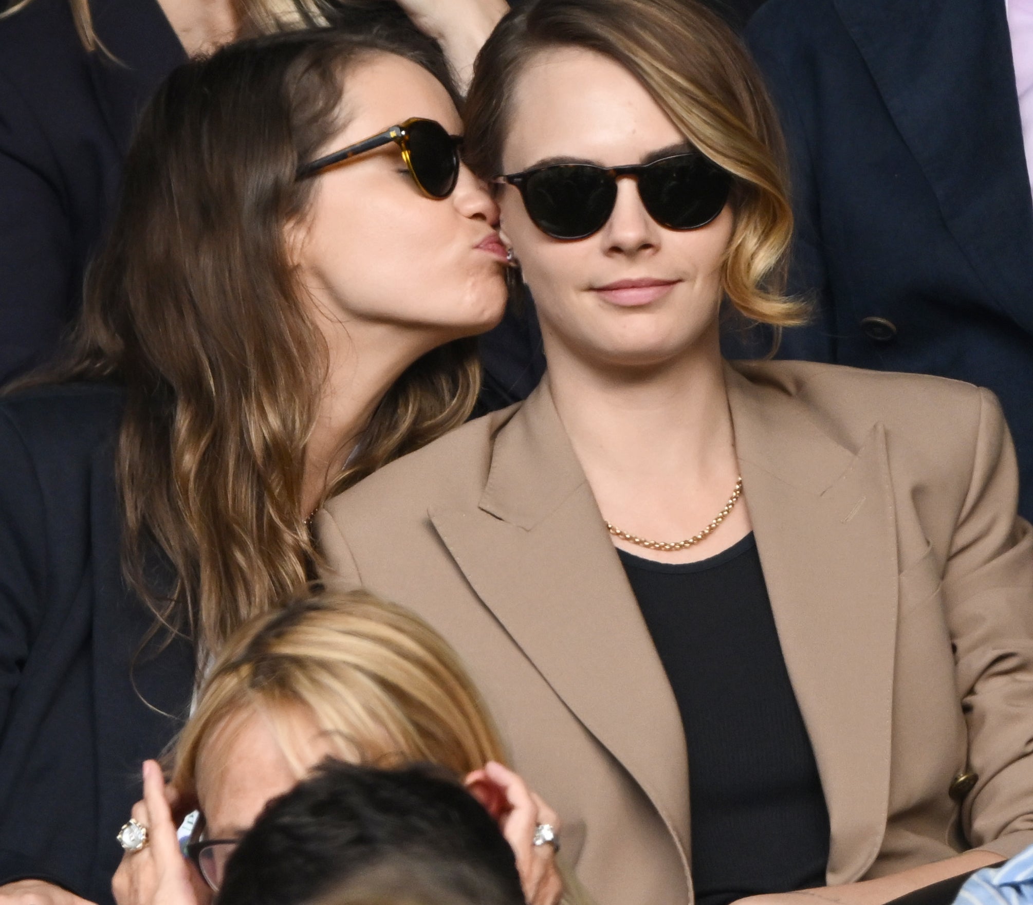 Minke kisses Cara Delevingne&#x27;s cheek as they sit in a crowded audience, both wearing sunglasses and stylish jackets