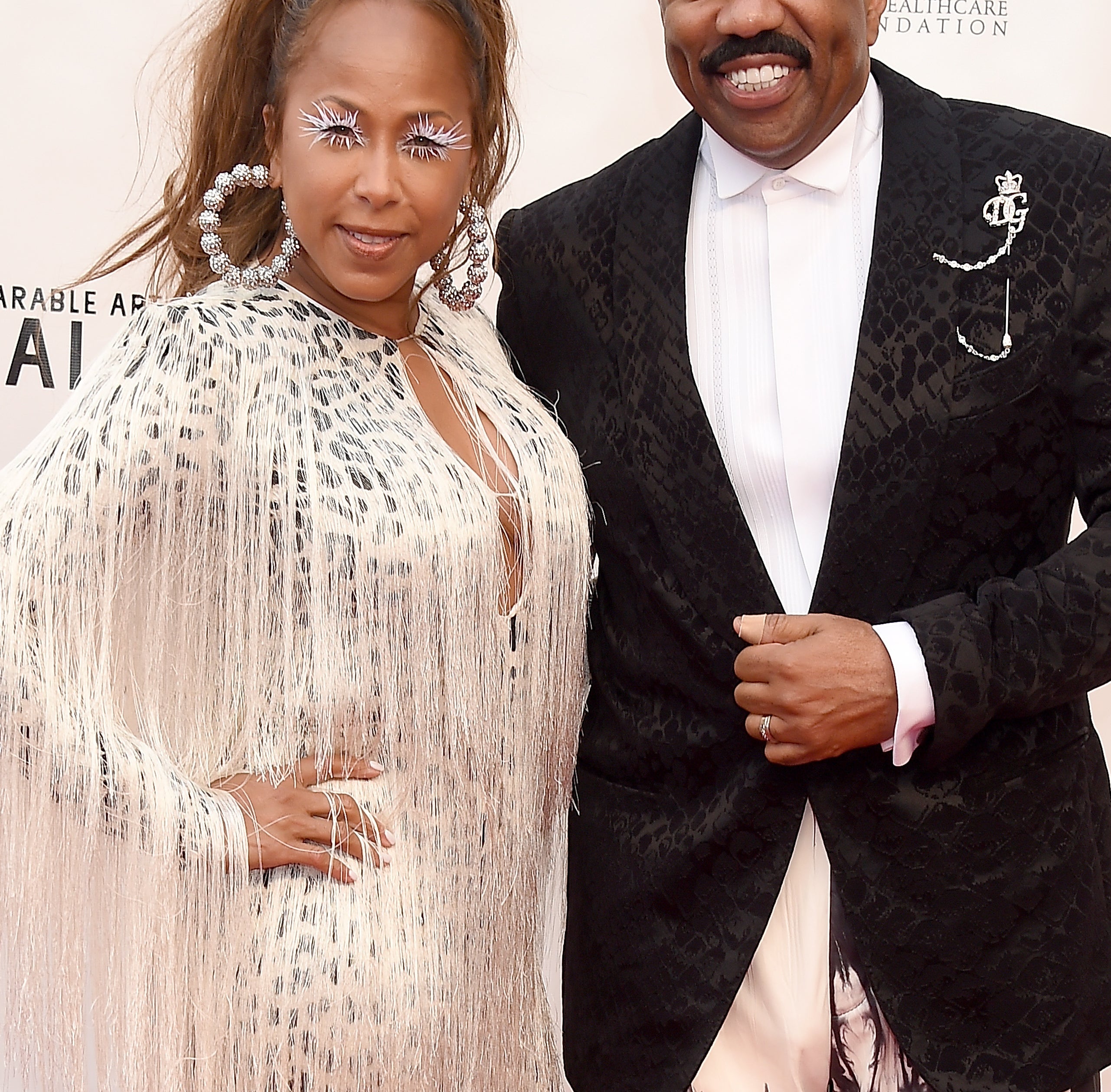 Steve Harvey and wife Marjorie on the red carpet at the Wearable Art Gala. Steve wears a black jacket and white pants; his guest wears a fringed, patterned dress