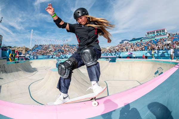 A young skateboarder in full protective gear performs a trick on a ramp at a skateboarding event in front of a large crowd