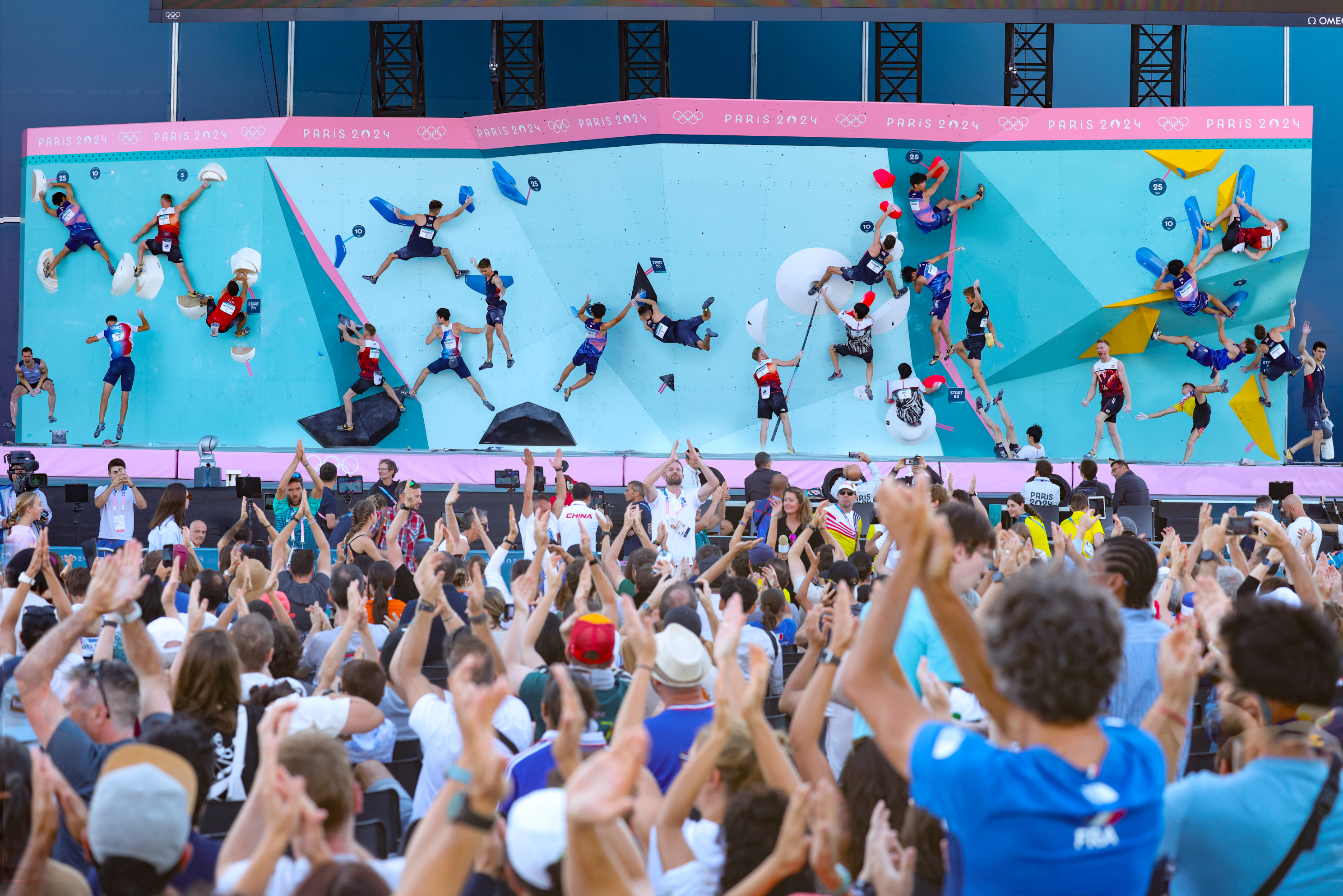 Crowd cheers in front of a climbing competition with multiple participants scaling walls on an outdoor stage