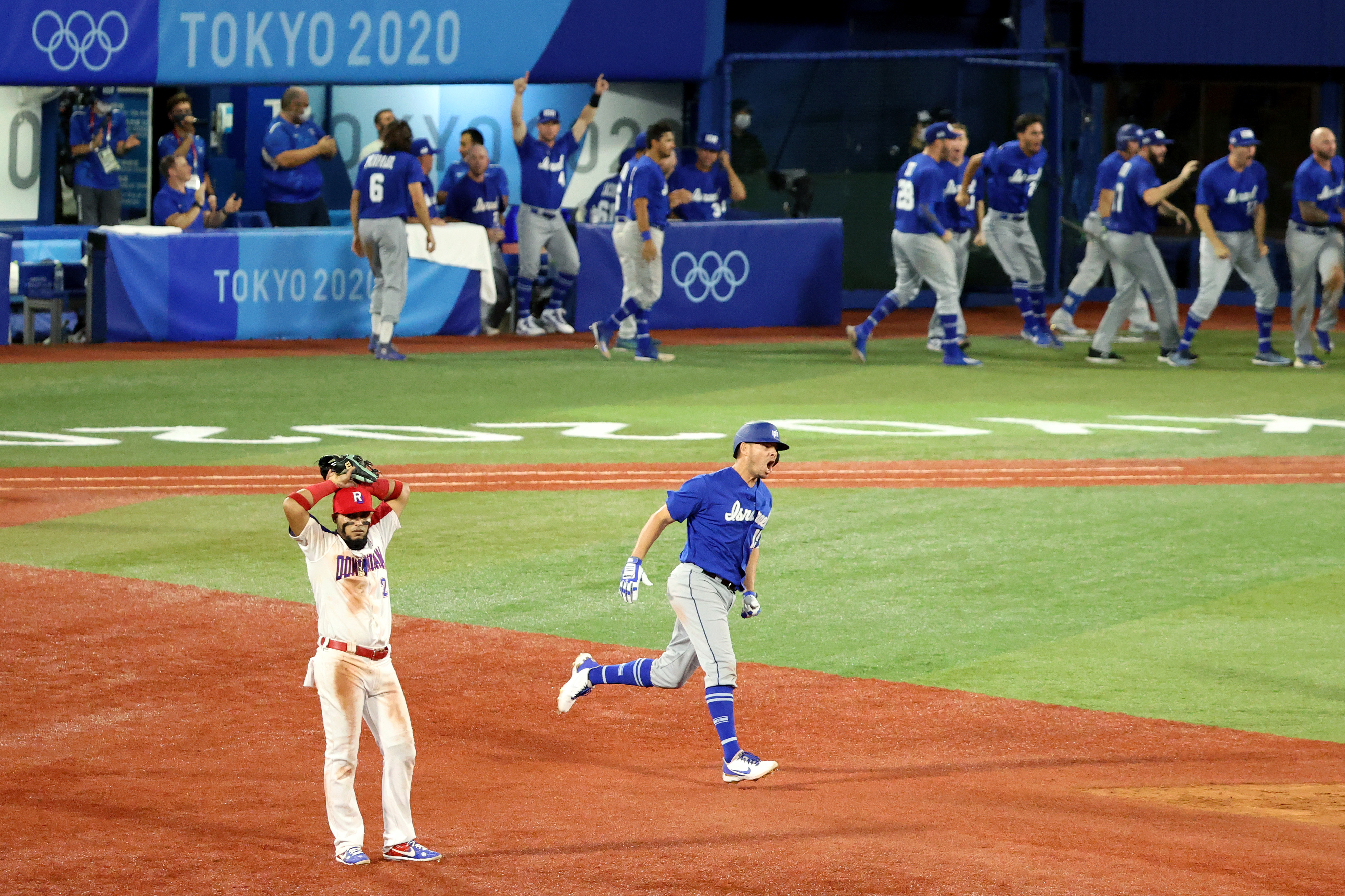 Baseball game at Tokyo 2020 Olympics: Dominican Republic player reacts as Israel player runs to home base with teammates celebrating in the background