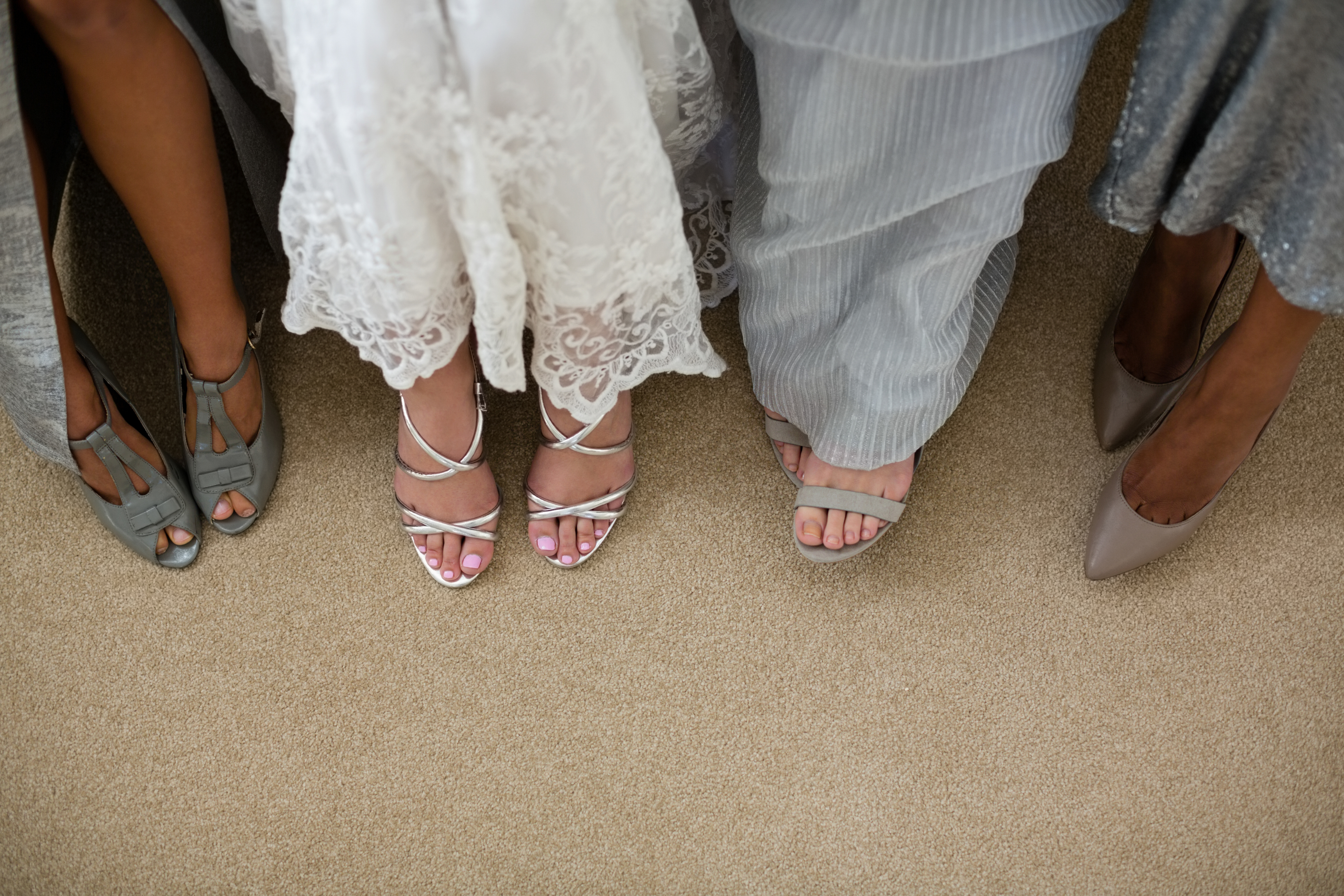 A close-up of three people’s feet in formal footwear. The middle person wears lace wedding attire with strappy heels. The other two wear matching dress shoes