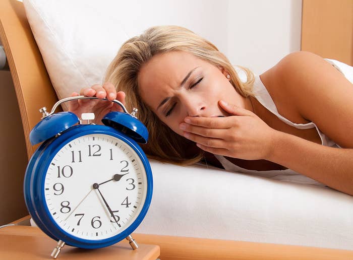 A woman lying in bed, yawning, and reaching out to turn off an old-fashioned alarm clock showing 7:00 am