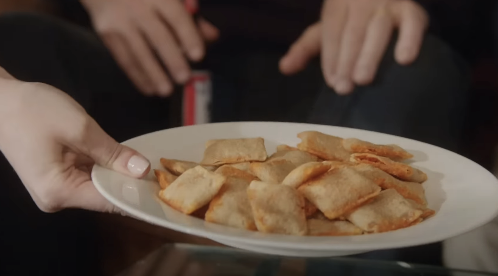 A person&#x27;s hand holds a plate of pizza rolls, with two people sitting in the background