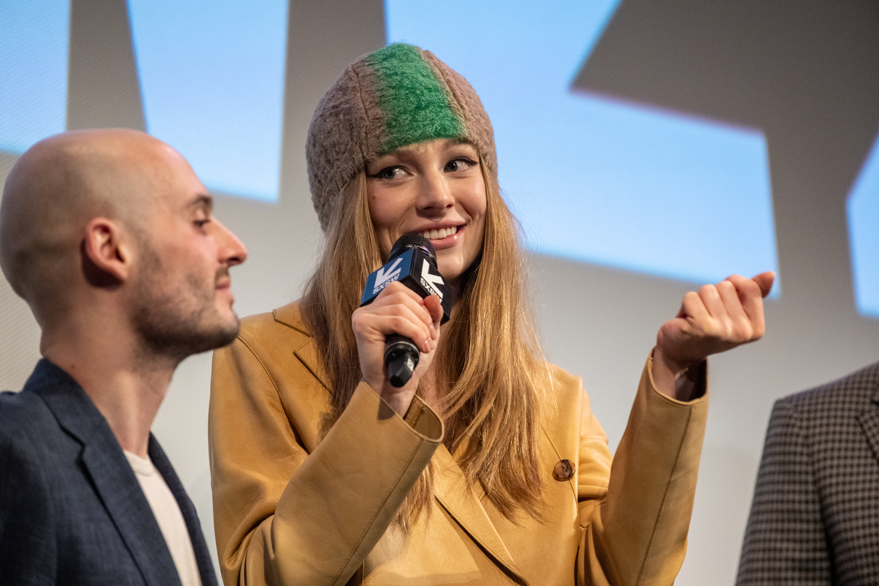 A man and Hunter Schafer, wearing a knitted hat and a yellow jacket, speaking into a microphone at an event