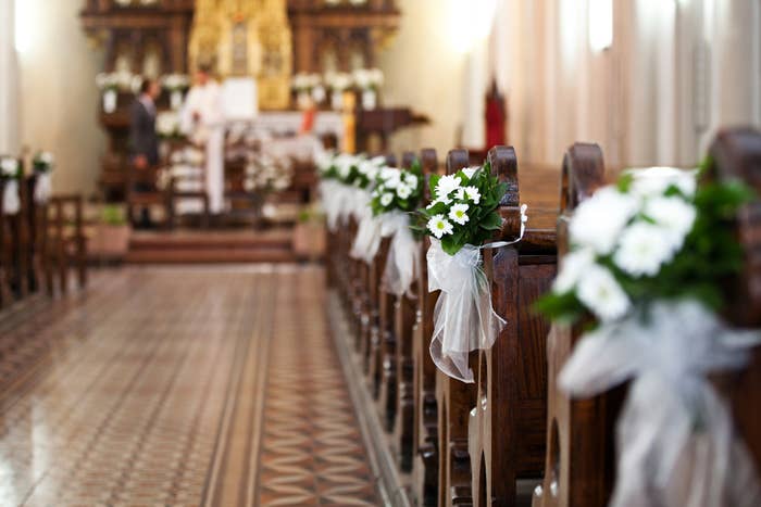 Church aisle decorated with white flowers and ribbons, leading up to an altar with a priest and another person in the background