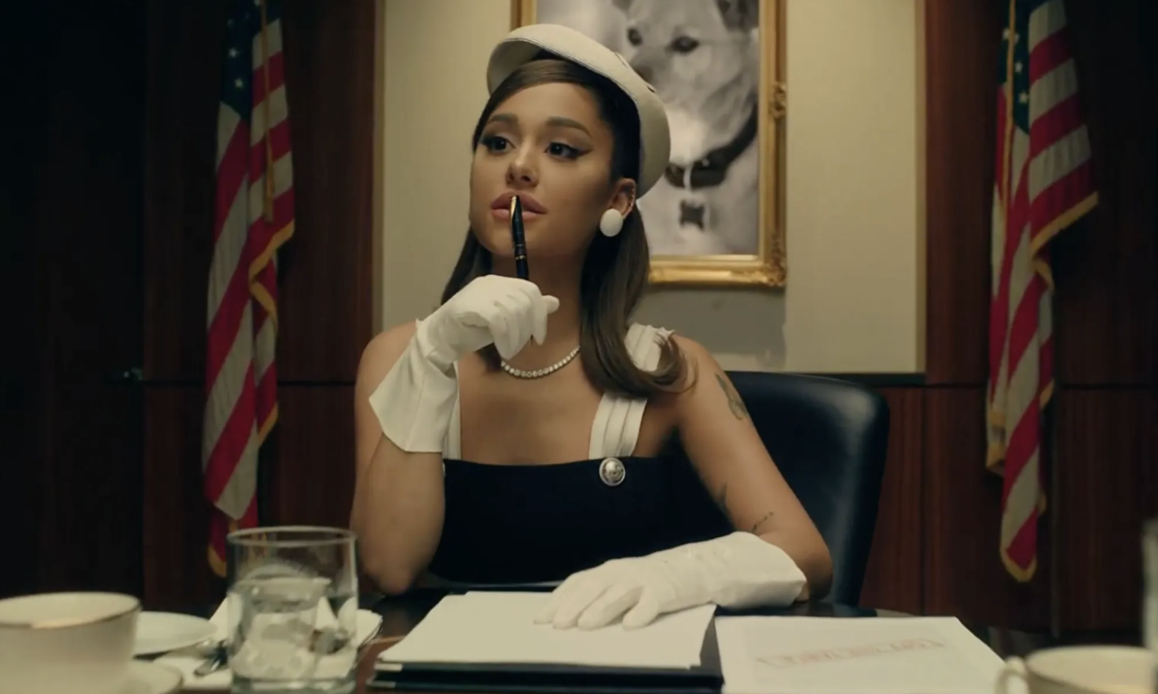 Ariana Grande sits at a desk in a classic, elegant outfit with white gloves and a pearl necklace, holding a pen to her lips in a thoughtful pose. Two flags and a dog portrait are behind her