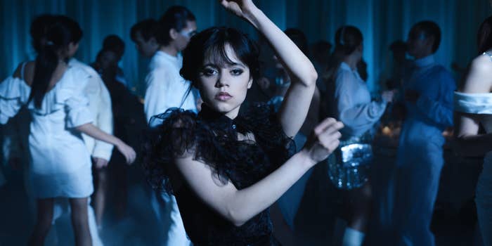 Jenna Ortega dances in a crowded room with people wearing formal attire. She wears a black ruffled dress