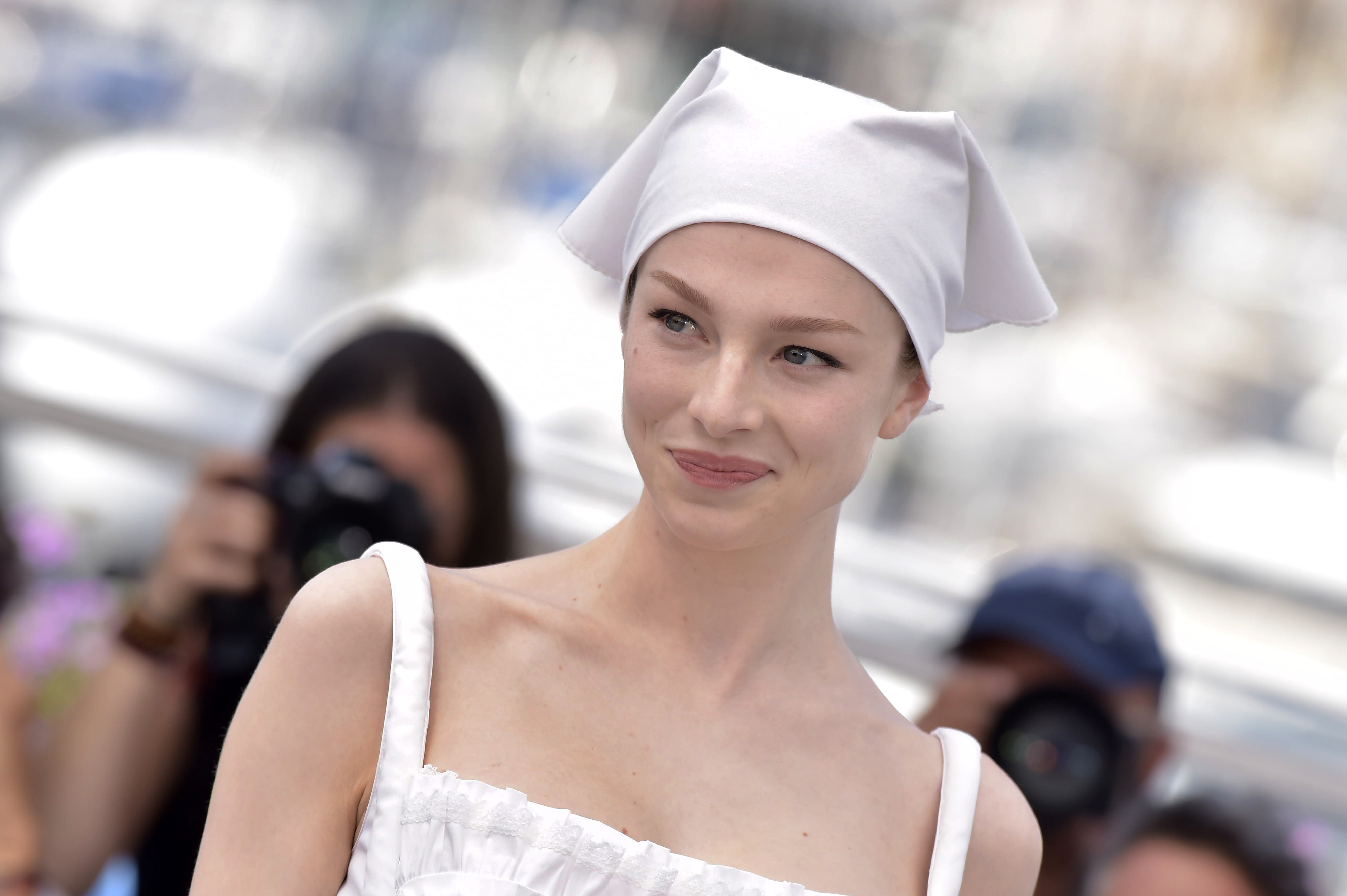 Emma Corrin on a red carpet, wearing a white dress with a matching headscarf. Photographers are visible in the background