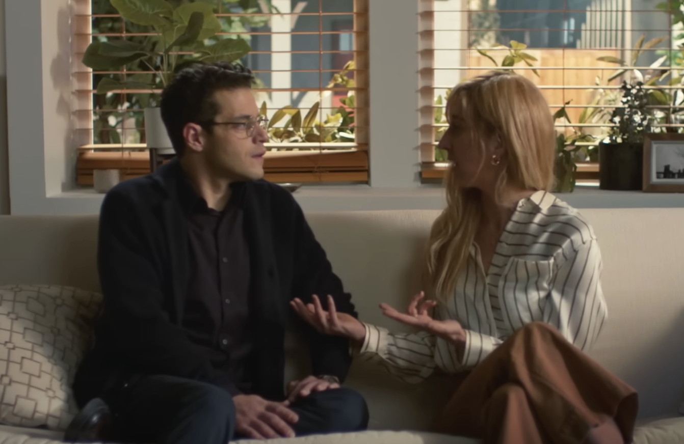 A man and a woman sit on a couch in a living room, engaged in a serious conversation. The man wears glasses and a dark sweater, while the woman has long hair and a striped blouse