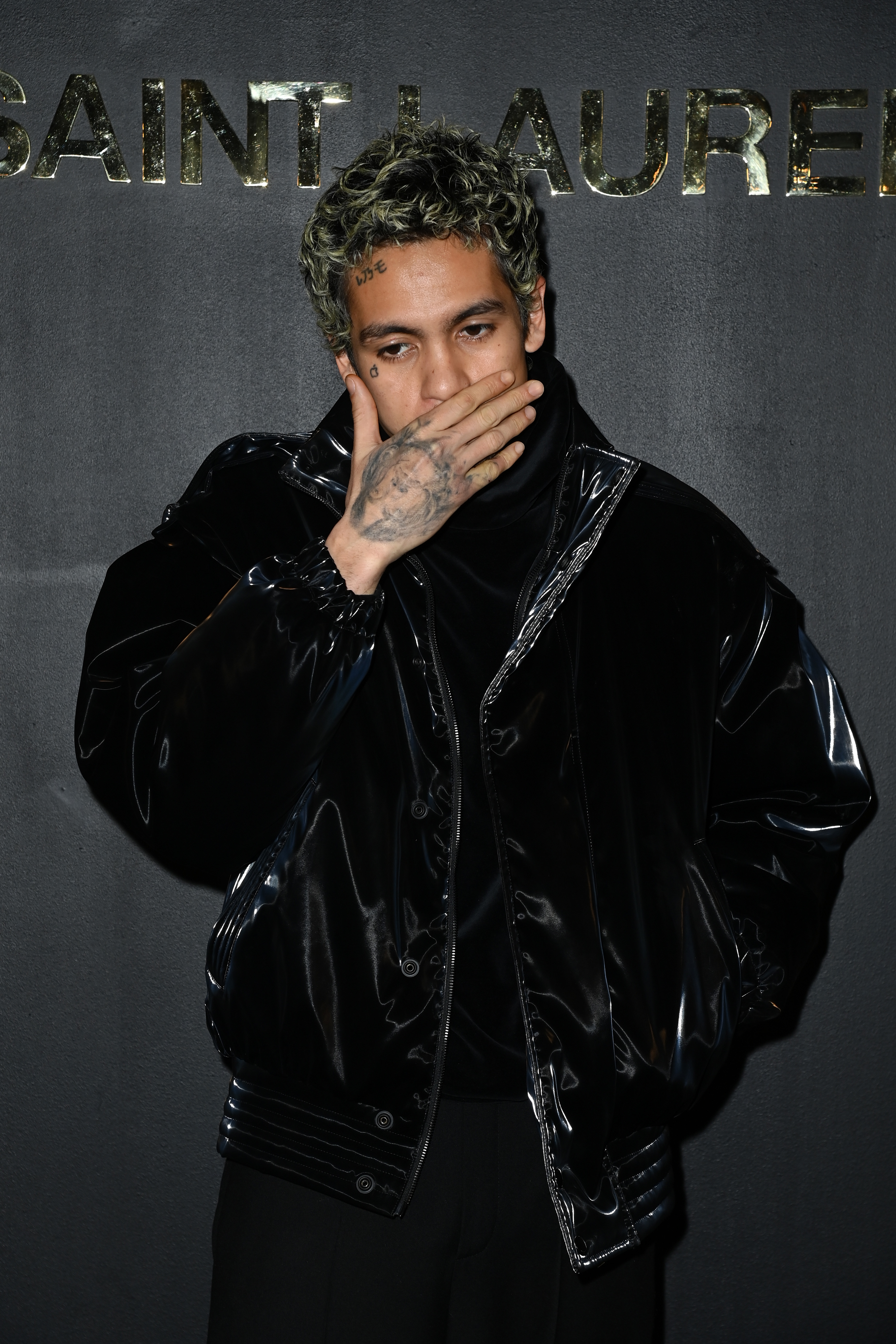 Dominic Fike, dressed in a shiny black jacket, poses with one hand covering his mouth against a backdrop with "Saint Laurent" written on it