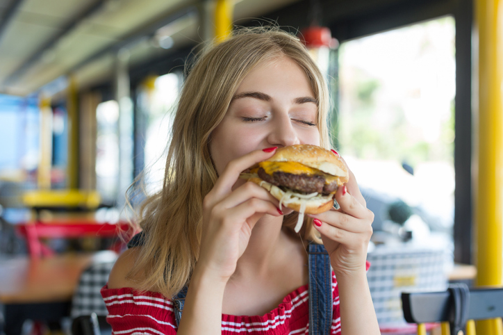 A woman with long hair, wearing a striped shirt, enjoys a bite of a cheeseburger in a casual dining setting