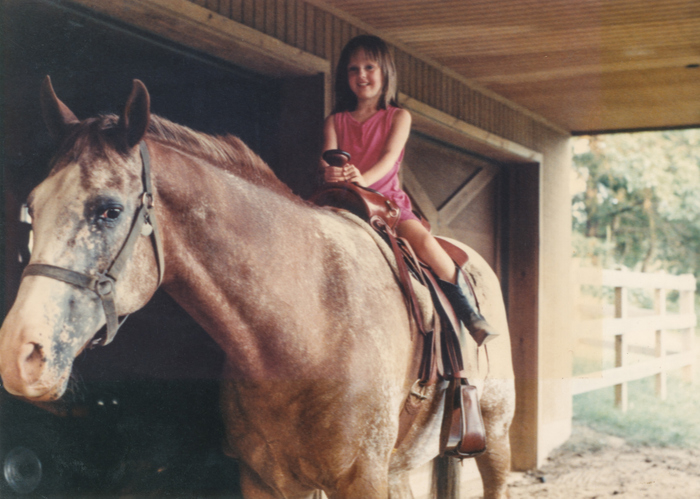 A young girl in a sleeveless dress sits on a horse inside a barn, smiling at the camera