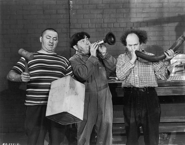 Curly Howard, Moe Howard, and Larry Fine of the Three Stooges play comically with musical instruments and a box, in a black-and-white photo