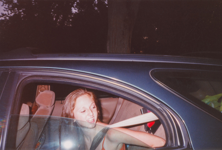 A person sitting in a car, smiling, and wearing a seatbelt, appears to be mid-conversation. The scene is informal and candid
