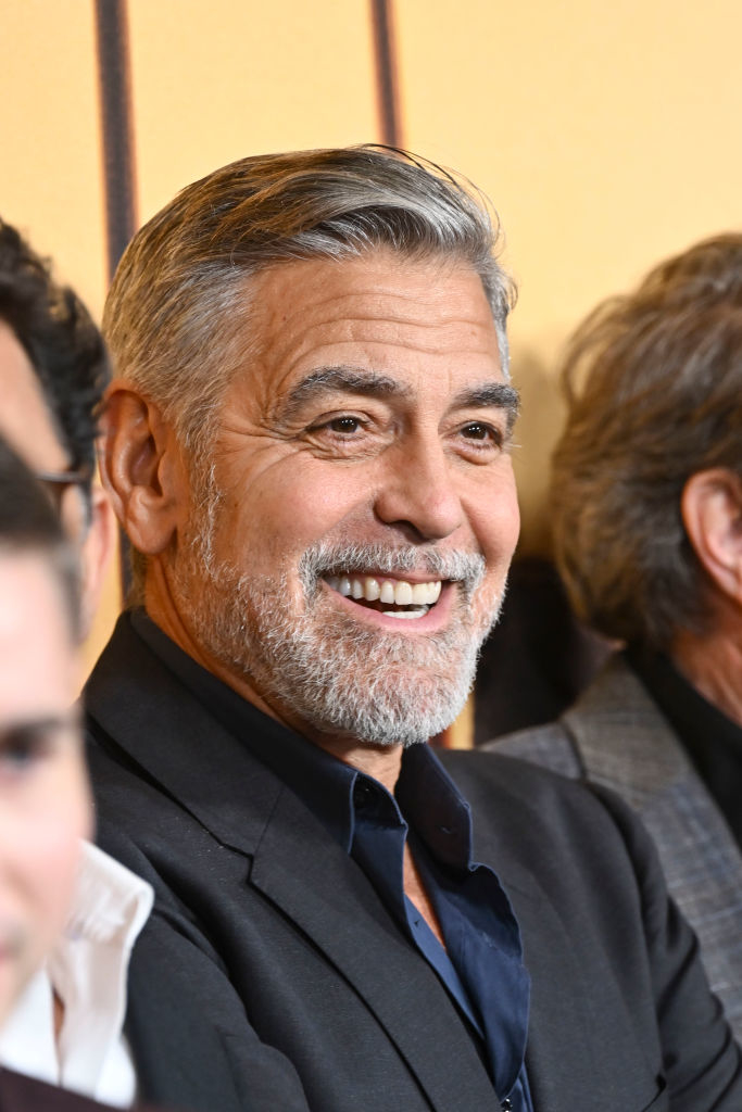 George Clooney smiling, wearing a dark suit and shirt, attending an event