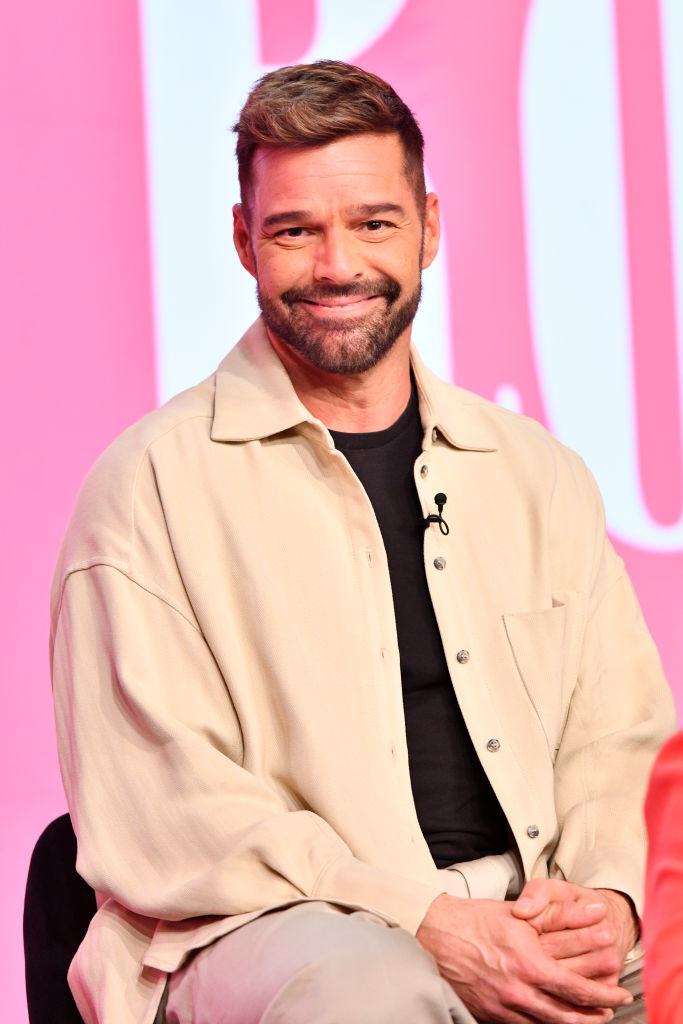 Ricky Martin smiling and sitting, wearing a beige shirt over a black top at a celebrity event