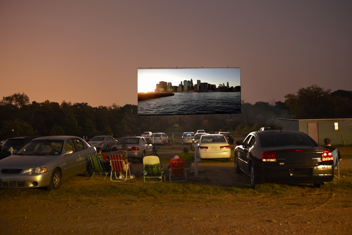Drive-in theatre at dusk with parked cars and people in lawn chairs watching a movie featuring a city skyline at sunset on a large outdoor screen