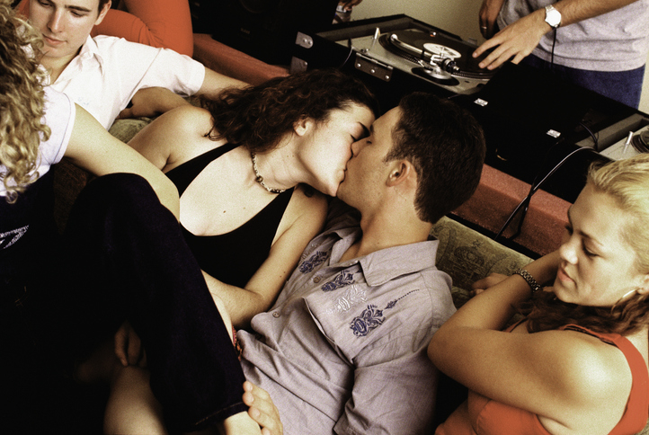 Four people at a party, two kissing on a sofa, while another person DJs in the background