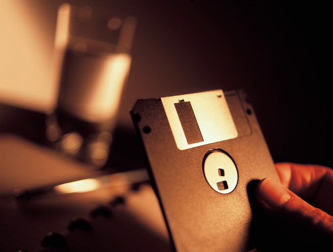 A hand holding a black floppy disk with a blurred glass of water in the background