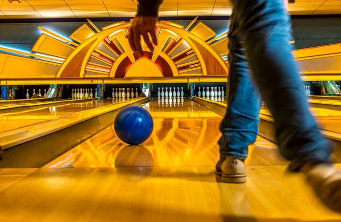 Person bowling, seen from behind, about to release a blue bowling ball down a lane toward pins. The setting appears to be a decorated bowling alley