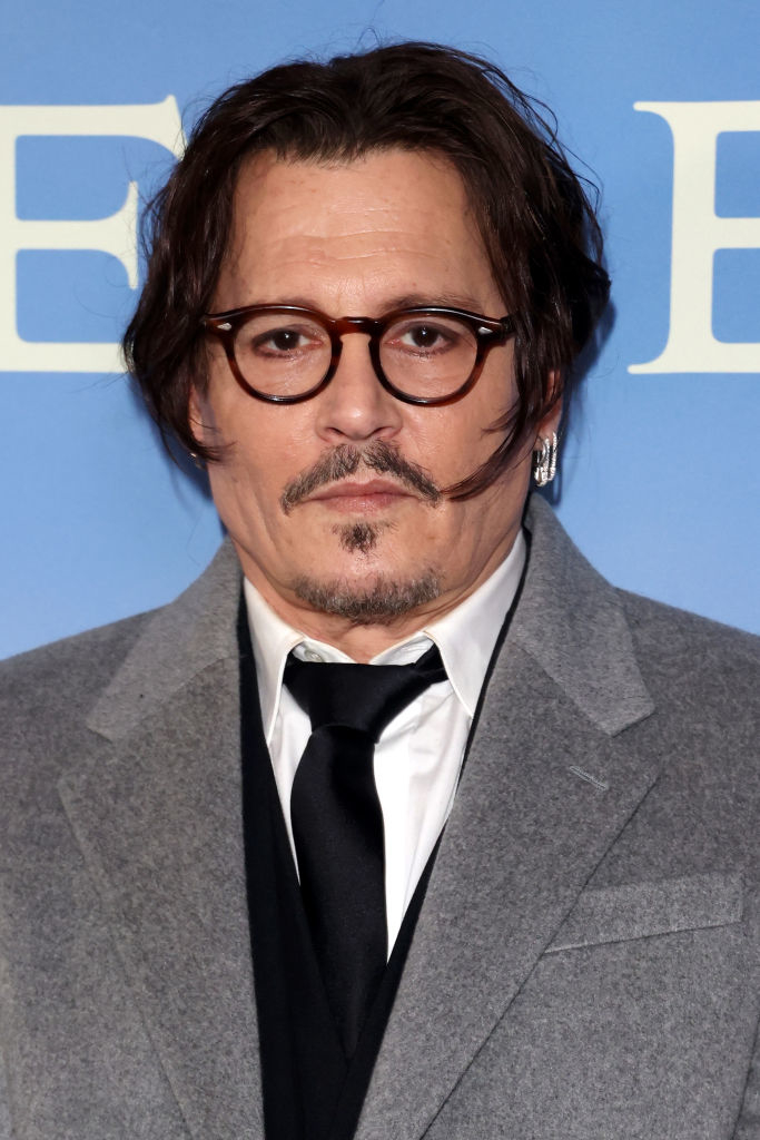 Johnny Depp poses in a suit with a shirt and dark tie, sporting round glasses, facial hair, and an earring, against a blue background