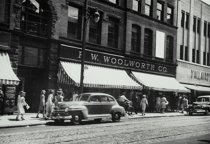 Black and white photo showing people walking in front of a Woolworth Co. store. 1940s cars are parked along the street, and striped awnings hang over the sidewalk