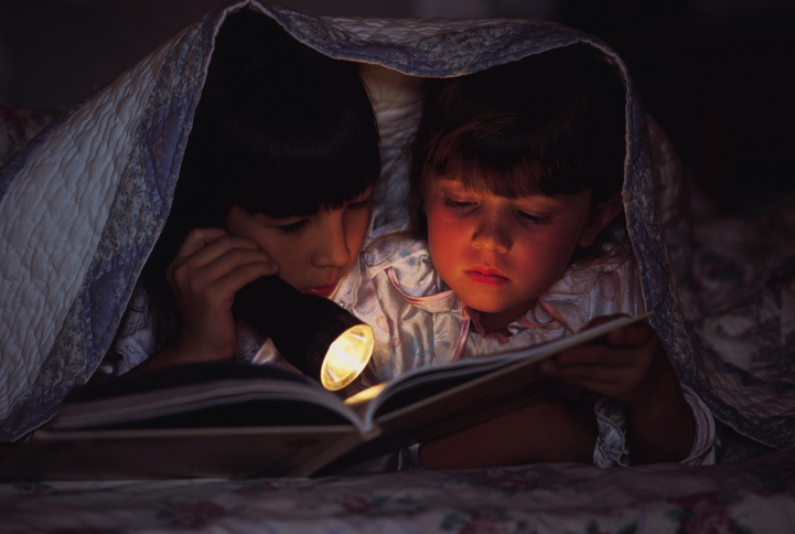 Two children read a book together under a blanket, illuminated by a flashlight