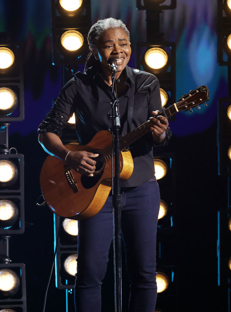 Tracy Chapman performing on stage with a guitar, dressed in a casual shirt and jeans, surrounded by bright stage lights