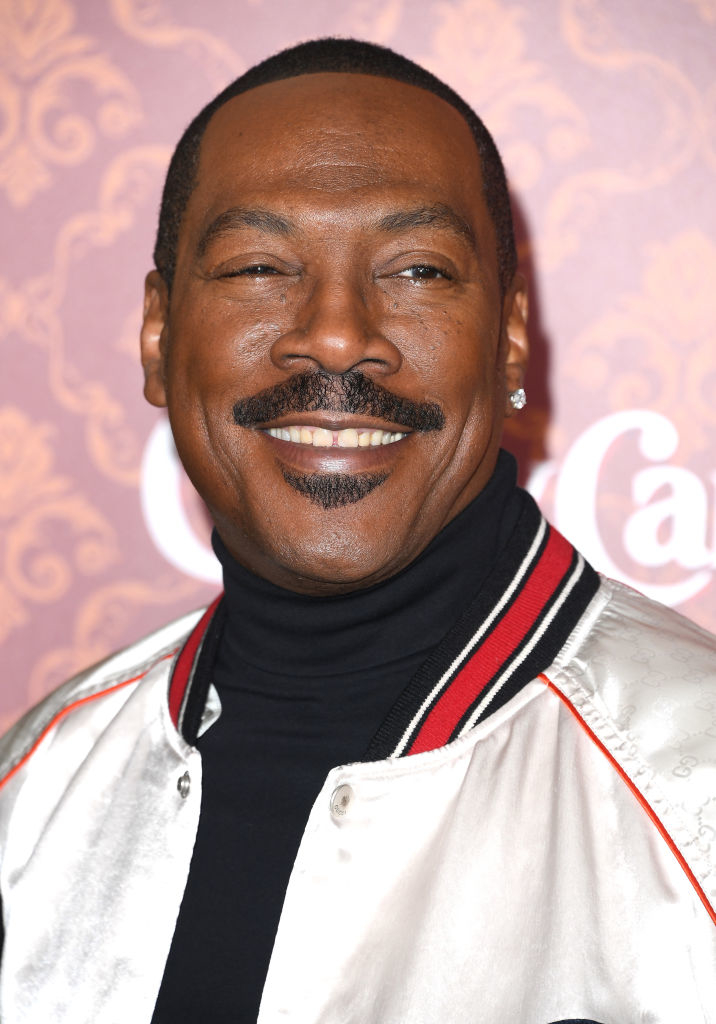 Eddie Murphy smiles while wearing a black turtleneck and a shiny jacket with red and black trim at an event