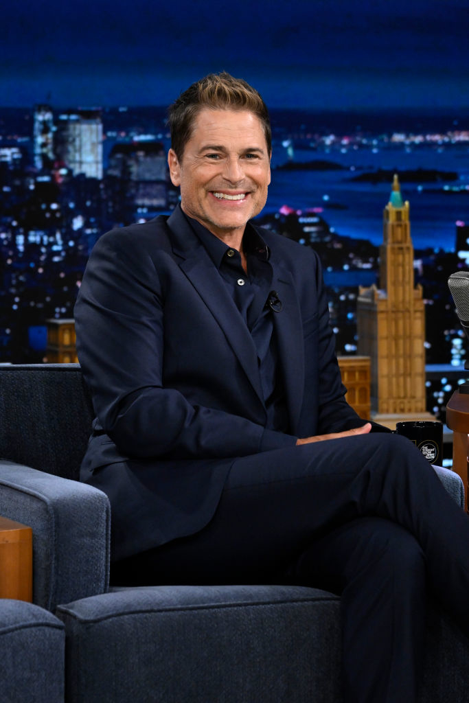 Rob Lowe wearing a dark suit, seated on a talk show set, smiles at the camera with a cityscape backdrop