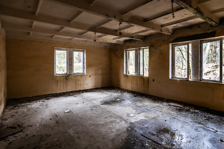 An empty, dilapidated room with peeling paint, broken windows, and debris scattered on the floor. The room appears abandoned and in disrepair