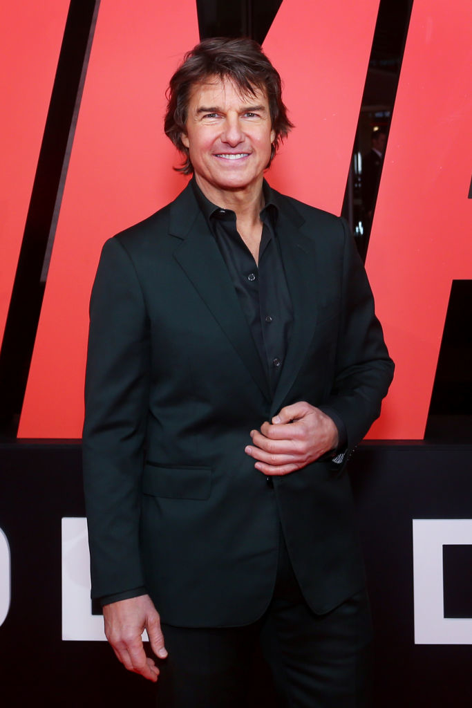 Tom Cruise on the red carpet wearing a black suit and black shirt, smiling at the camera