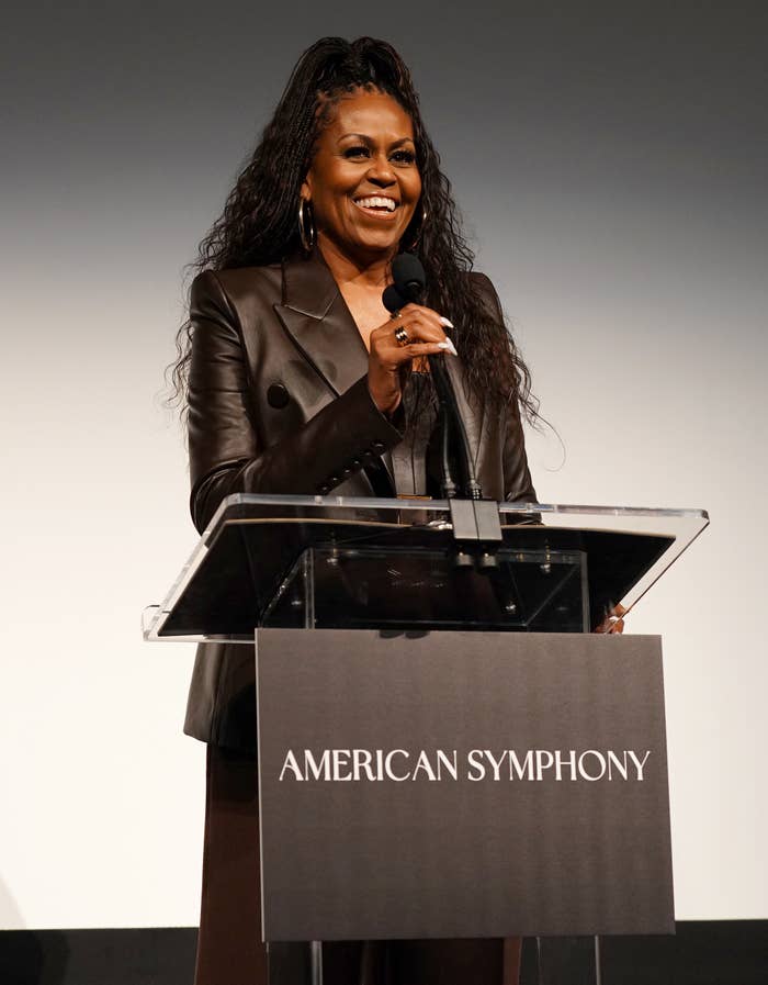 Michelle Obama smiling while speaking at a podium labeled "American Symphony," wearing a stylish dark suit