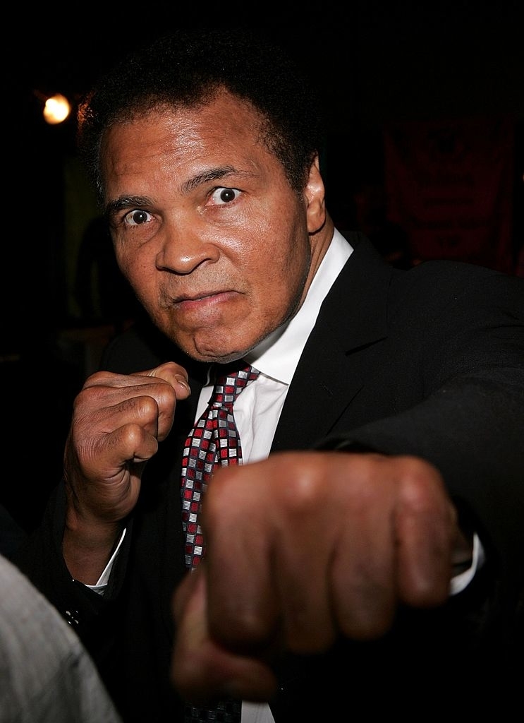 Muhammad Ali poses with a fist forward in a suit and patterned tie