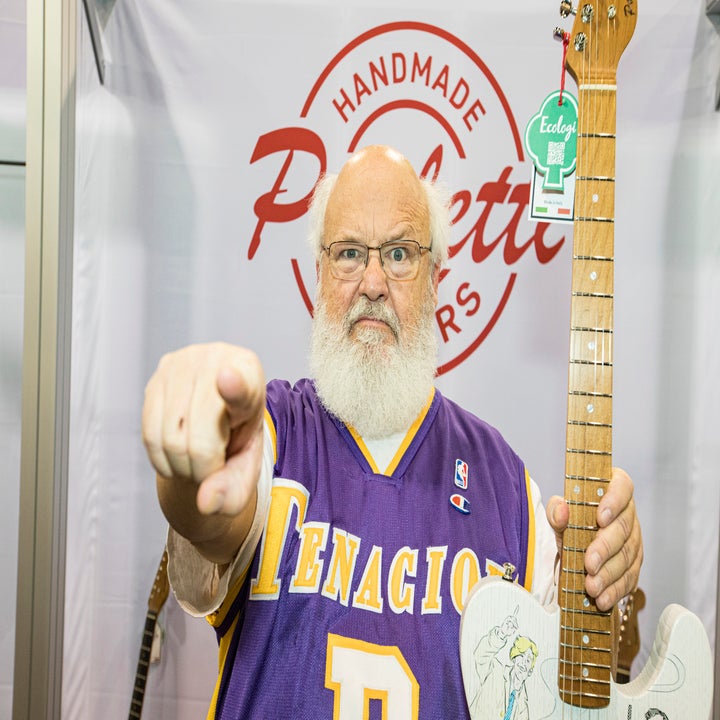 An older man with a white beard and glasses, wearing a "Tenacious D" basketball jersey, points at the camera while holding a guitar