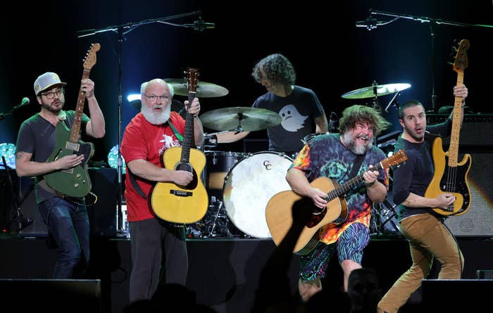 Kyle Gass, Jack Black, and bandmates perform on stage with guitars and drums. Jack Black wears a tie-dye shirt and shorts, while Kyle Gass wears casual clothing