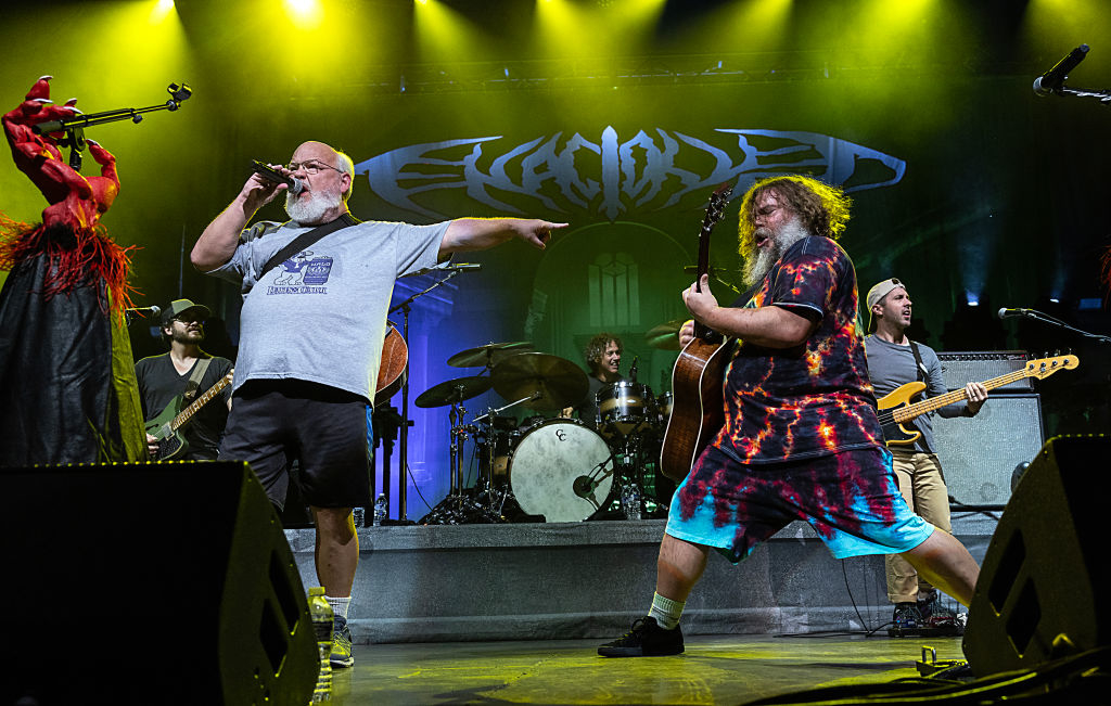 Jack Black and Kyle Gass of Tenacious D perform energetically on stage with their band at a concert, with a dramatic lighting setup in the background
