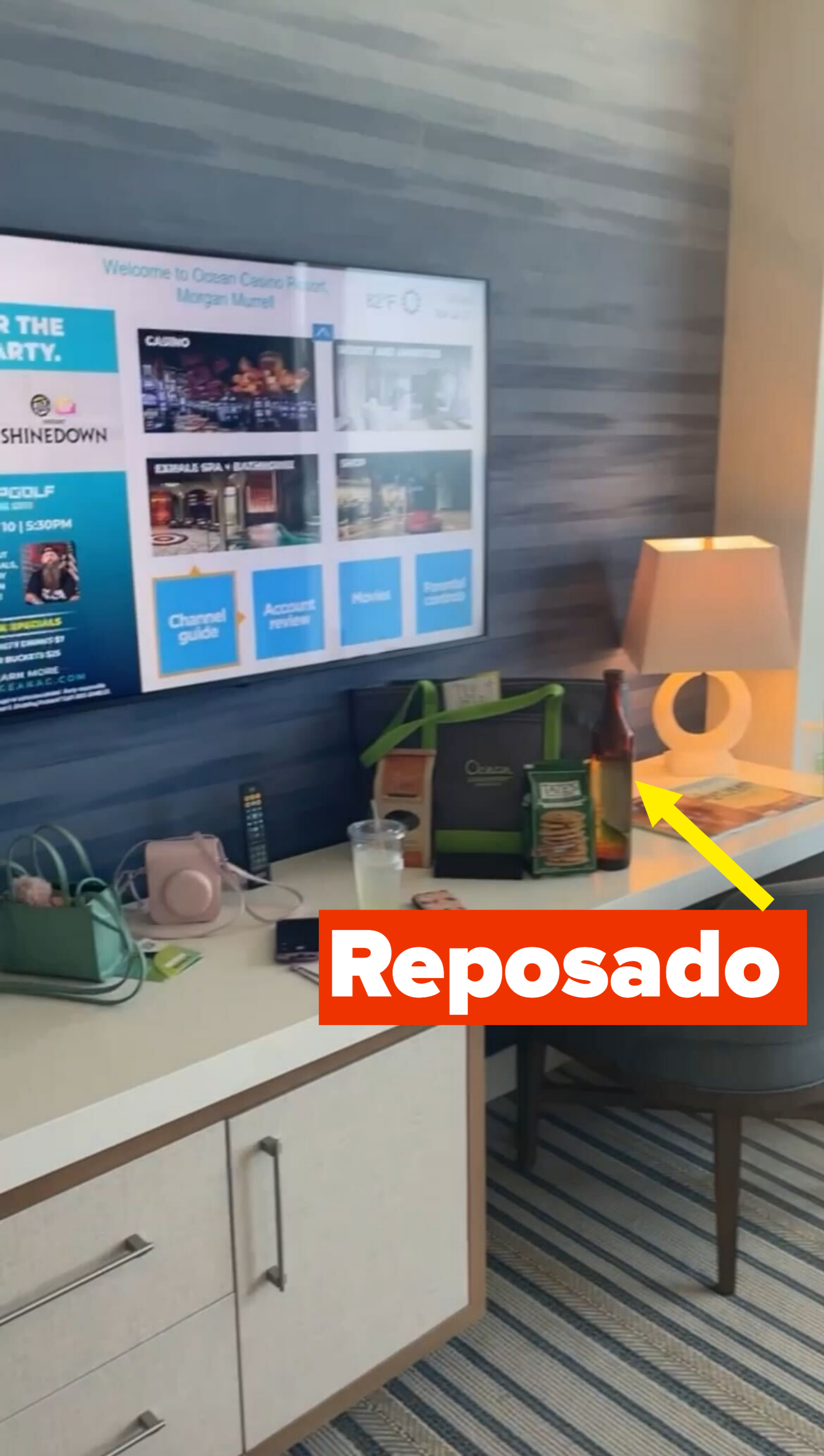 Hotel room desk area with a lit lamp, items on the desk include a green bag, water bottle, snacks, and a TV mounted on the wall with various displayed content