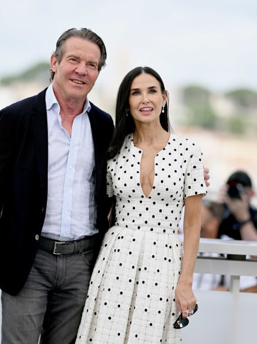 Dennis Quaid and Demi Moore pose together outdoors. Dennis wears a dark blazer and gray trousers, while Demi wears a white polka-dot dress with a deep neckline. Photographers are visible in the background