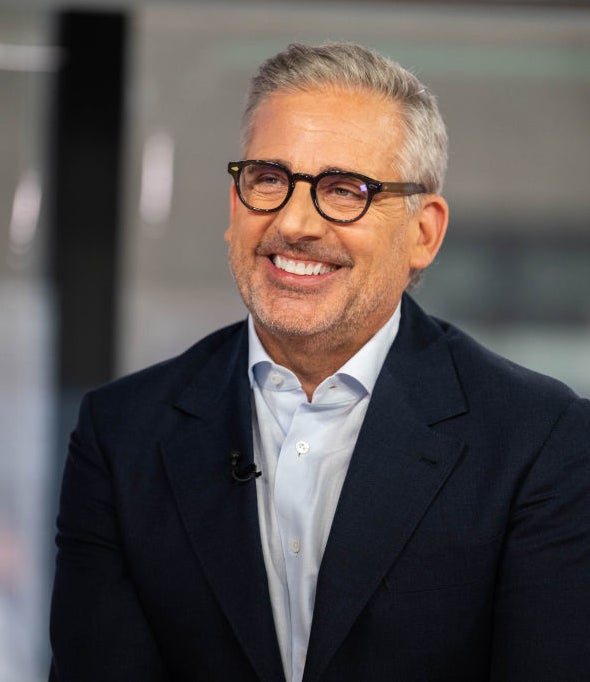 Steve Carell smiling in an interview setting, wearing glasses, a white shirt, and a dark blazer